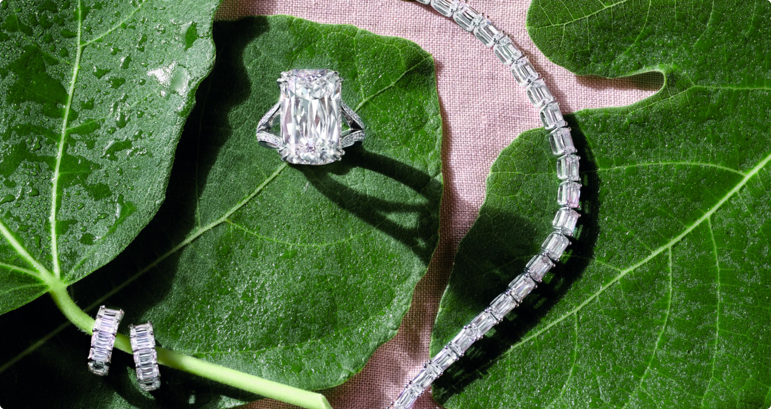 A luxurious engagement ring with a large emerald-cut diamond set on a silver band, alongside a diamond bracelet, both resting on dew-covered green leaves.