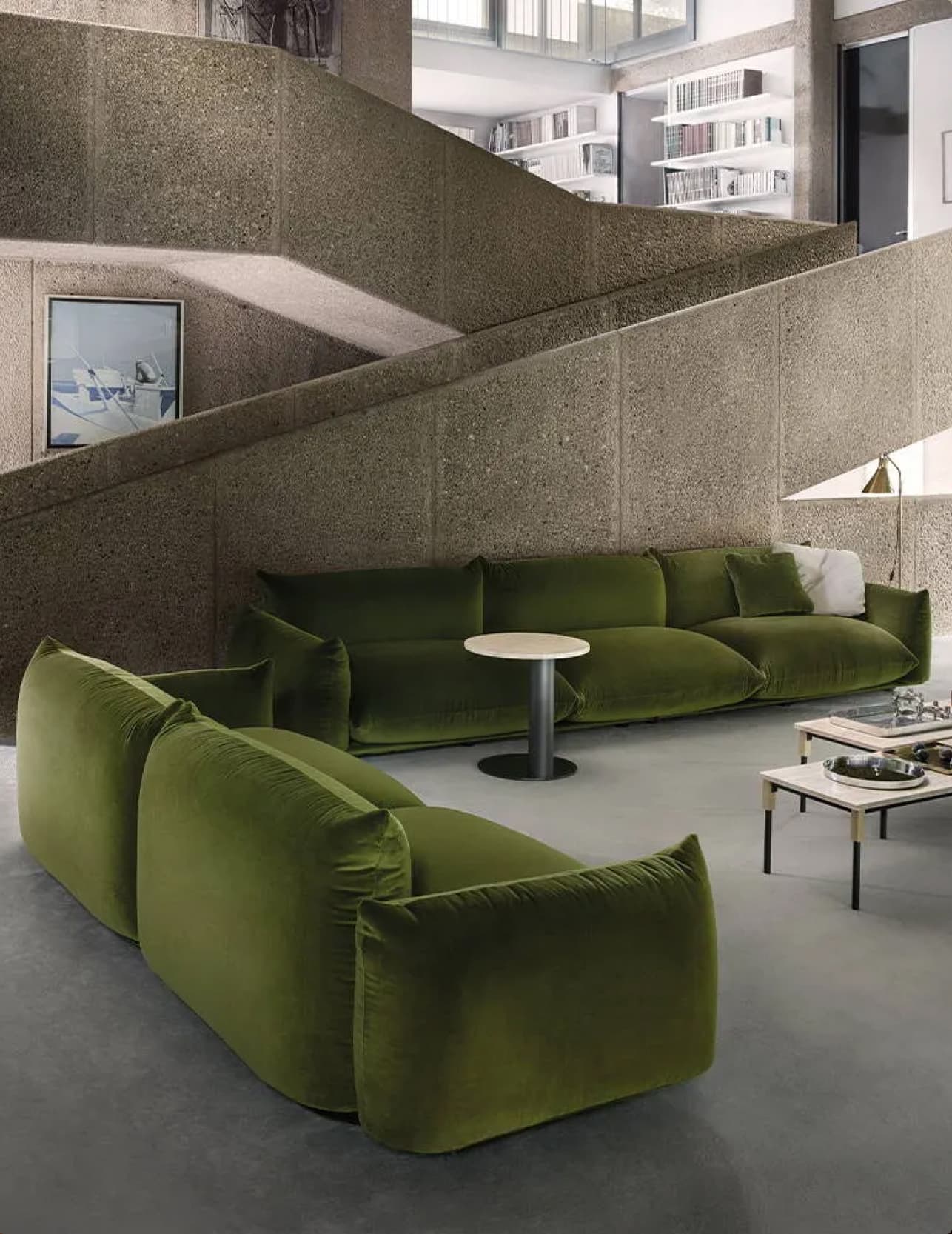 A stark, modern living space with a concrete stairwell. The focal point of the image are comfortable, contemporary green sofas arranged around various side tables.