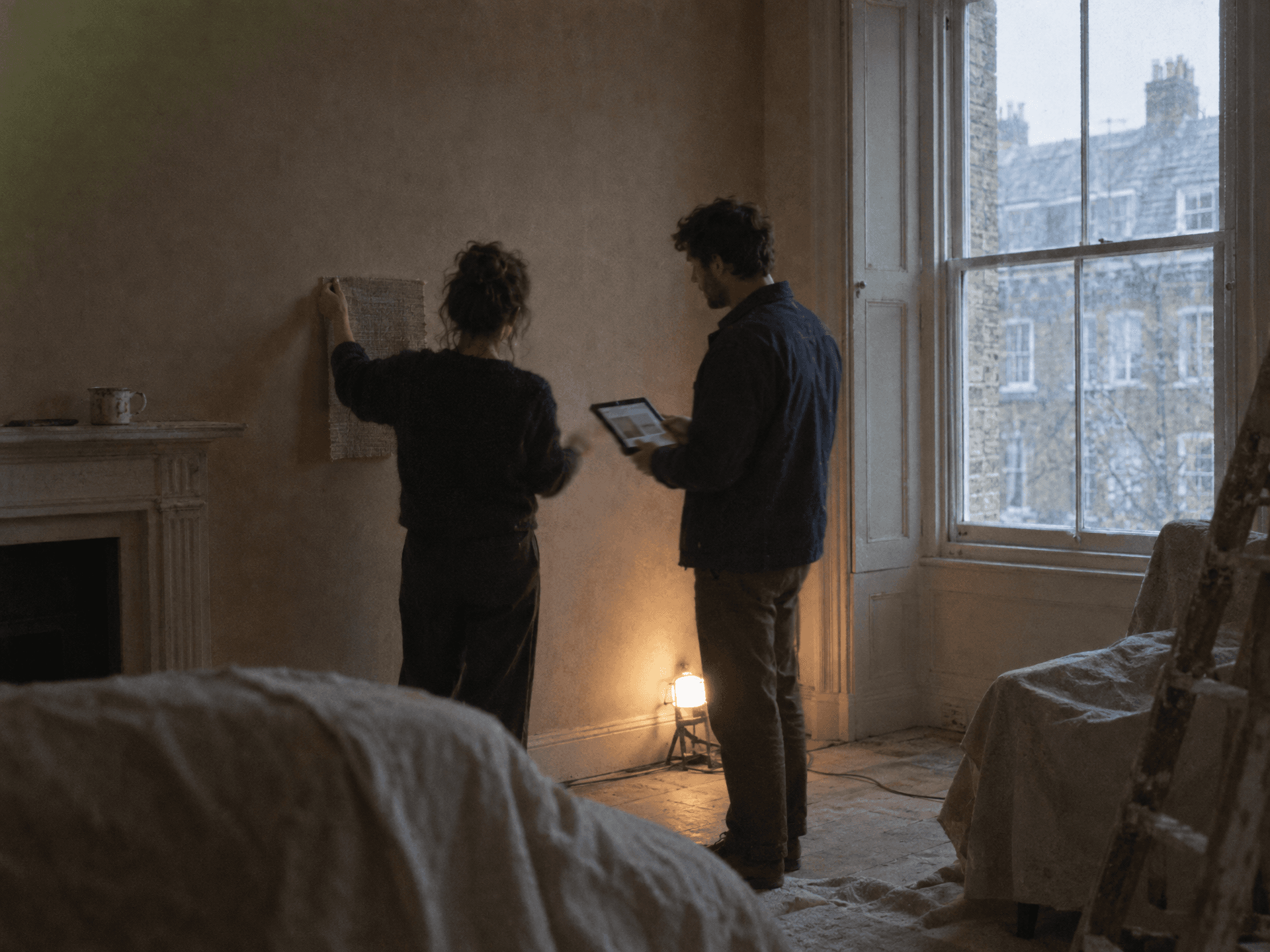 A woman holds a woven fabric sample up against a bare plaster wall while a man beside her looks at a tablet, the two comparing it in a period room mid-renovation. Dust sheets cover the furniture and a tall sash window looks onto a terrace of houses.