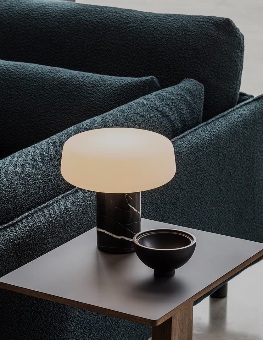 A modern table lamp with a black marble base and a white shade sits beside a black bowl on a wooden side table next to a dark textured sofa.